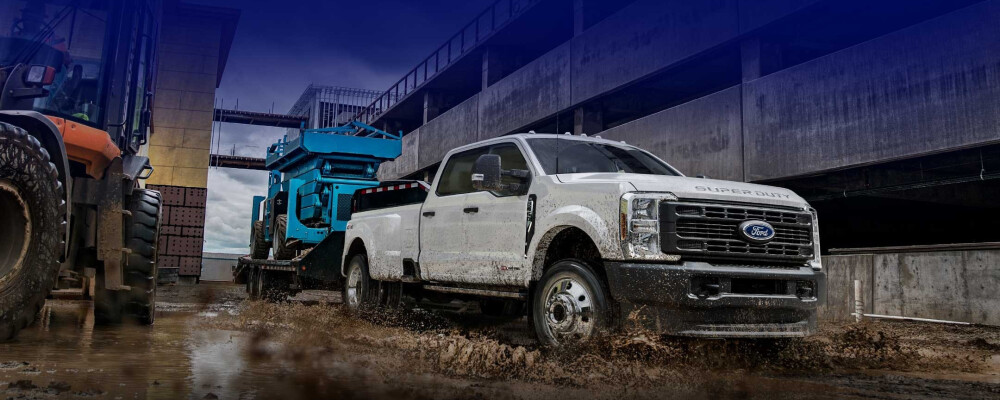 White Ford Super Duty truck towing large blue machinery through muddy construction site, conveying ruggedness and strength. Building framework visible in background.