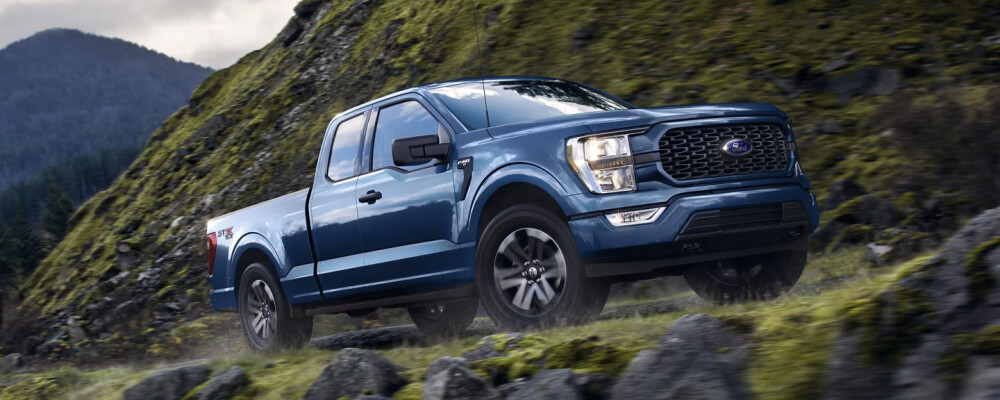 Blue pickup truck driving on a rocky, moss-covered hillside. The background features misty mountains and a cloudy sky, conveying a rugged adventure.
