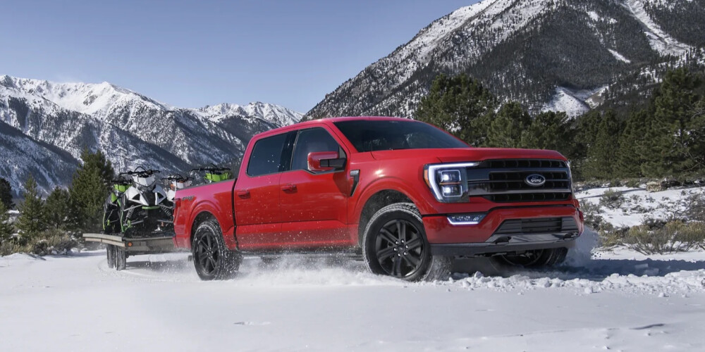 A red truck drives through snow, towing two snowmobiles on a trailer. Snowy mountains and pine trees are in the background under a clear blue sky.