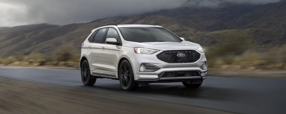 A silver SUV drives on a wet road with mountainous terrain in the background. The overcast sky adds a moody and adventurous tone to the scene.