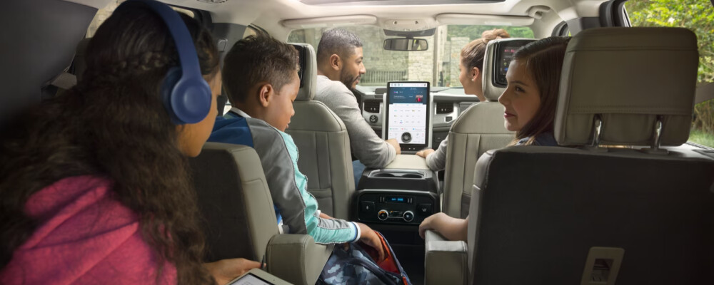 A family of five sits in a car, facing forward. The driver and passenger converse, while children in the back engage with devices. Mood is lively and interactive.