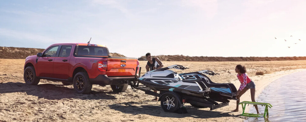 A red pickup truck parked on a sandy beach with two people unloading a jet ski near the water. The sun sets in the background, creating a relaxed atmosphere.