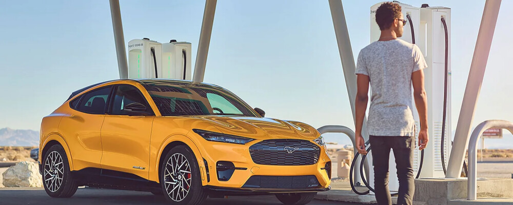 A man approaches an electric car charging station in an open landscape. A bright yellow car is parked beside him, under a clear blue sky, suggesting innovation.