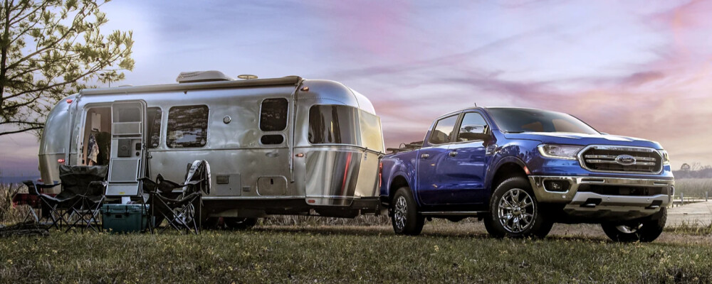 A blue pickup truck is parked beside a shiny silver camper on a grassy area at sunset. Camping chairs and a cooler are set up nearby, creating a relaxed, adventurous vibe.