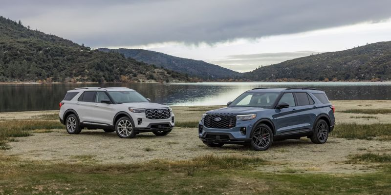 Two SUVs parked by a tranquil lake, surrounded by lush green hills under a cloudy sky. The scene conveys a sense of adventure and calm.
