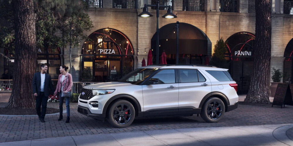 A white SUV is parked on a cobblestone street near a pizza restaurant. A couple walks past, smiling and talking. The setting is urban and relaxed.