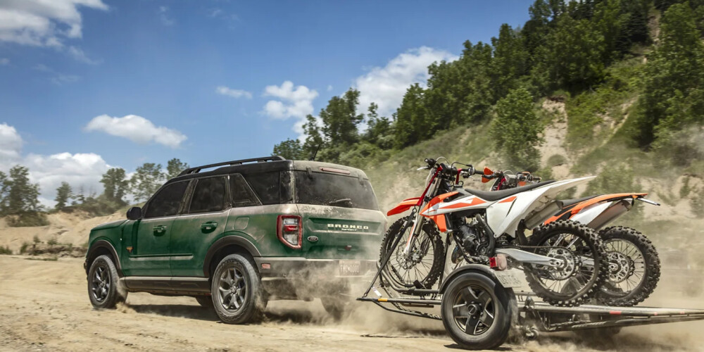 A green SUV drives on a dusty road, towing a trailer with two dirt bikes. The scene is set under a blue sky with clouds, surrounded by lush trees, conveying adventure.