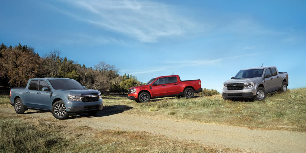 Three pickup trucks are parked on a grassy hill under a clear blue sky. From left to right, there's a grey, a red, and another grey truck. The scene is peaceful and open.